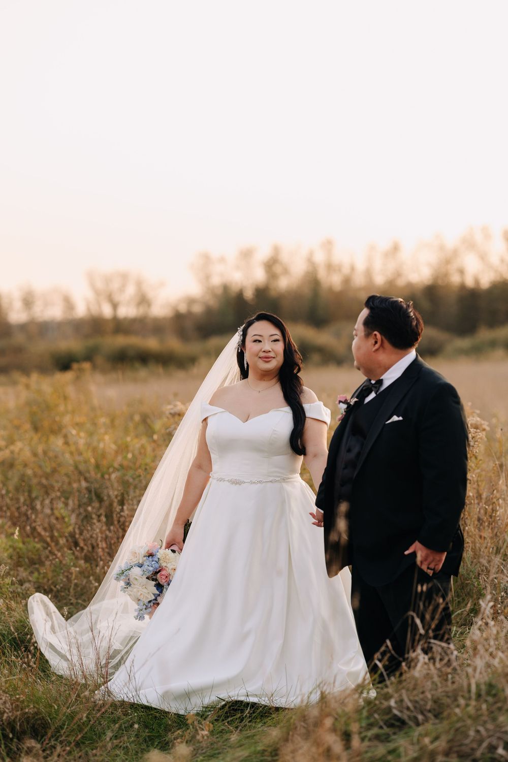 Bride in white gown and groom in black suit sit together in autumn field.
