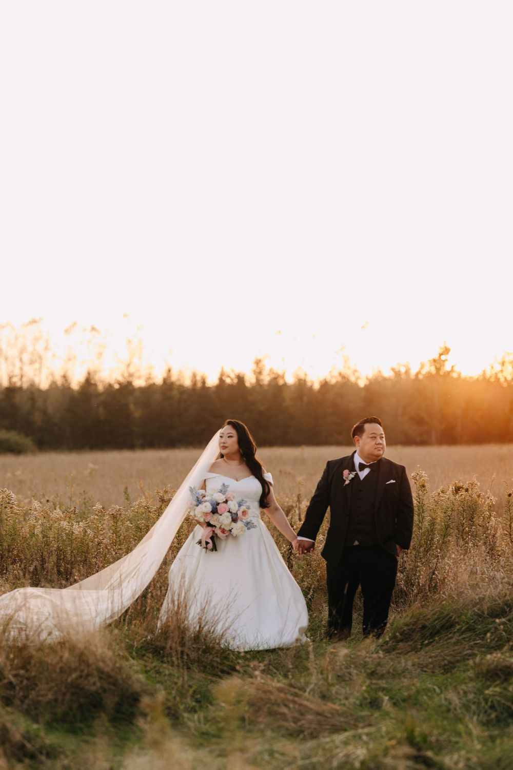 Couple takes romantic wedding photos in field with golden sunset behind trees.