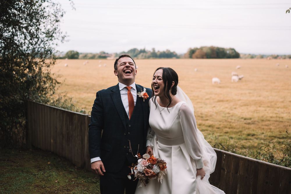 A joyful couple laughs together on their wedding day in front of a rustic field landscape.