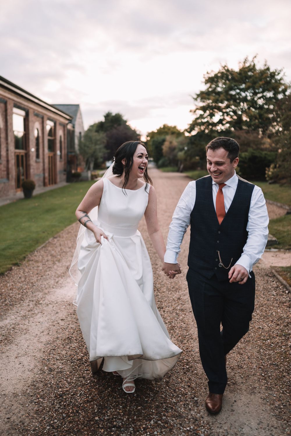 A couple walking together outdoors on a gravel path, with bride in white dress and groom in dark vest and orange tie.