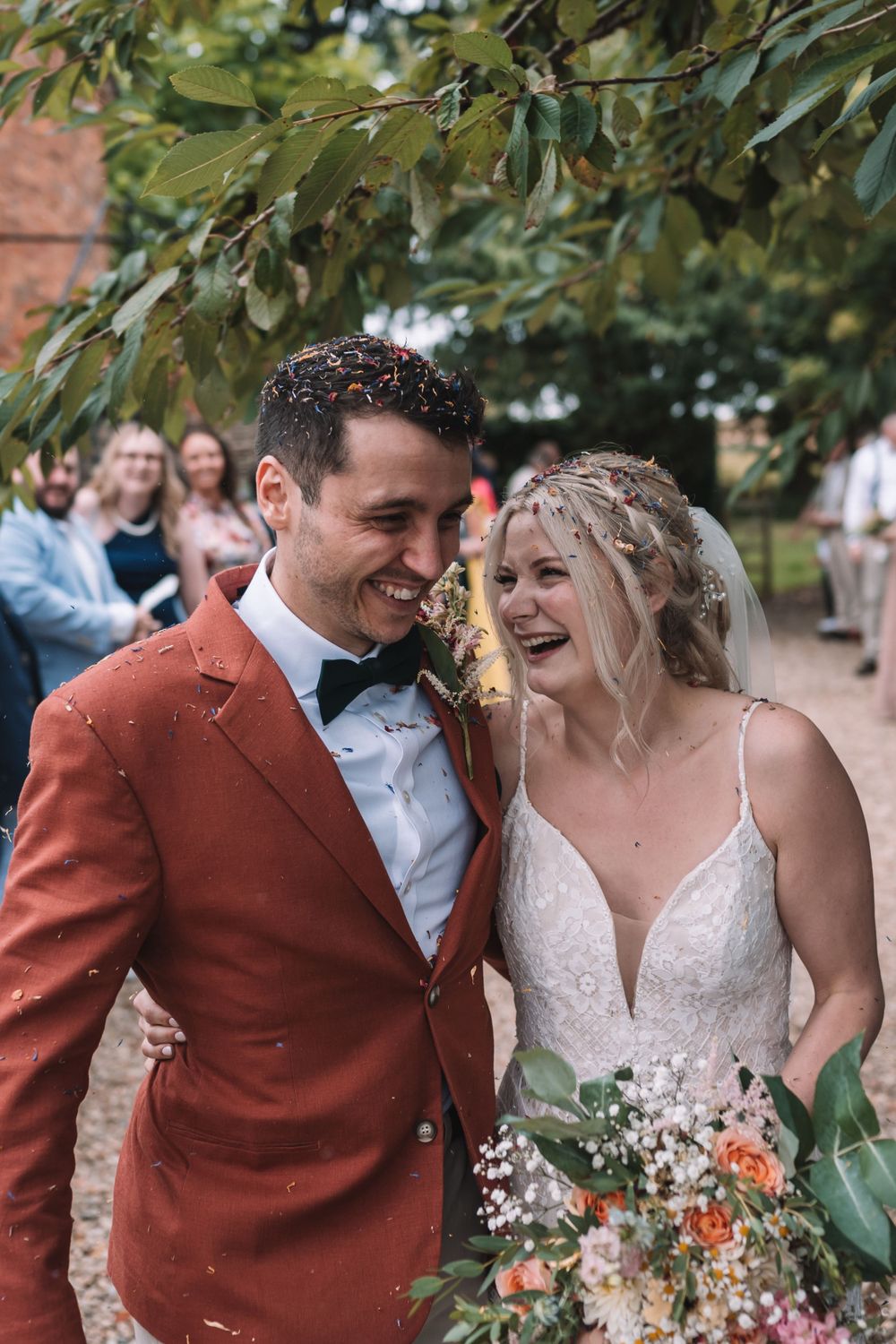 A couple in wedding attire sharing a joyful moment, with the groom wearing a rust-colored suit and bride in white lace dress.