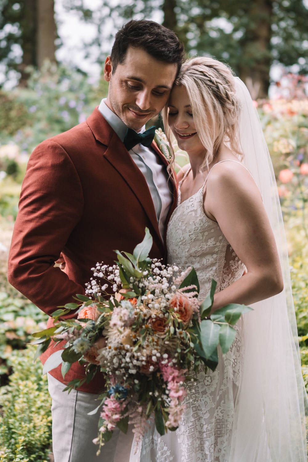 A romantic outdoor wedding portrait features a bride in lace and groom in burgundy jacket with a wild flower bouquet.