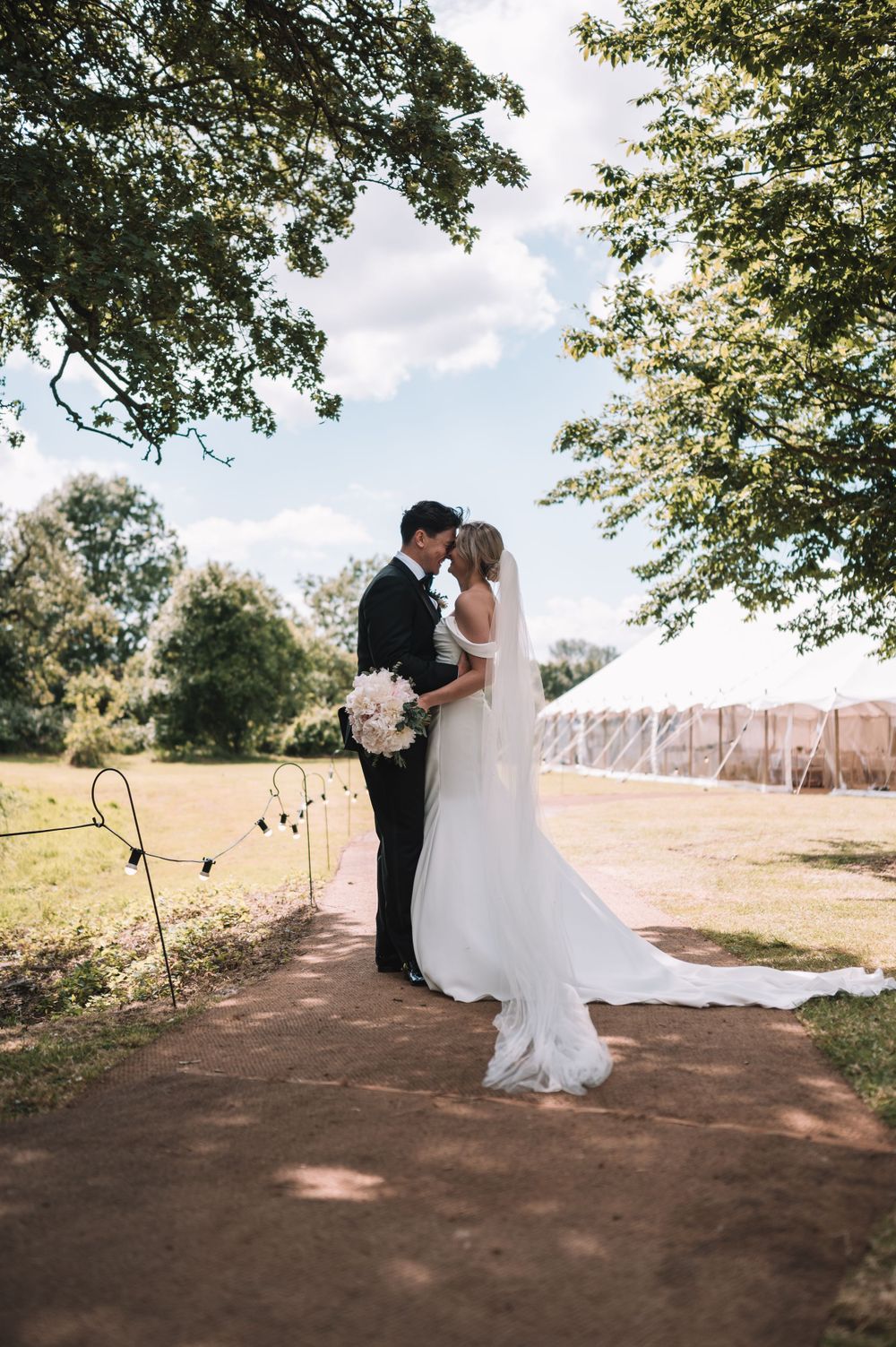 Romantic outdoor wedding scene on a sunny day with trees and string lights in a rustic country setting.