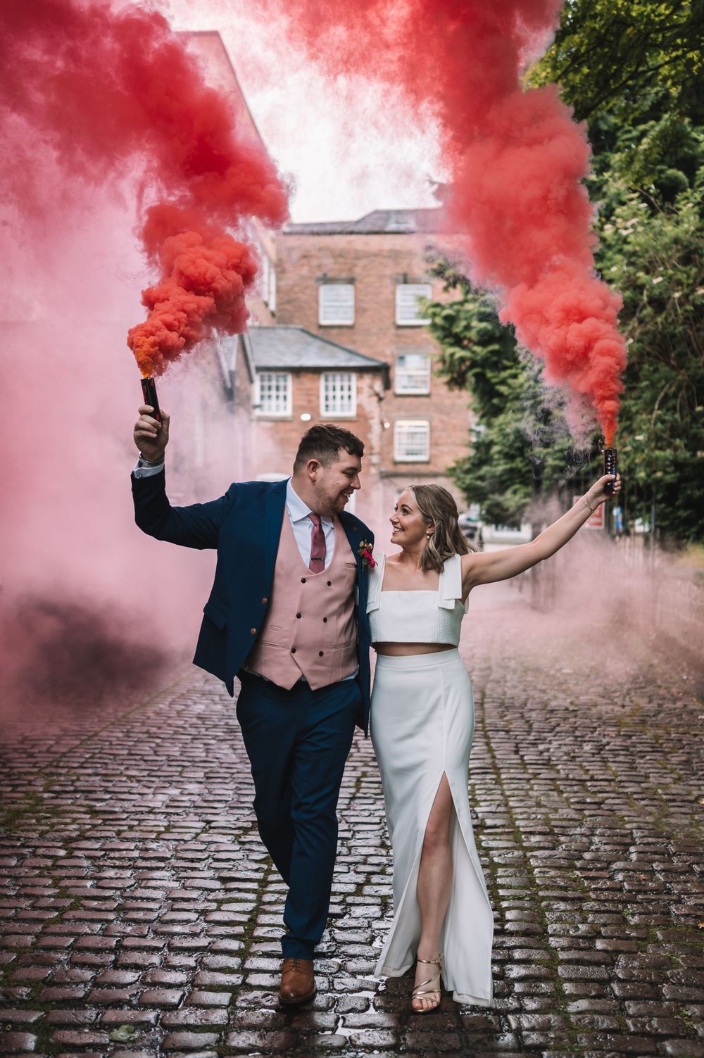 Couple celebrates on cobblestone street with red smoke flares in front of historic brick building.