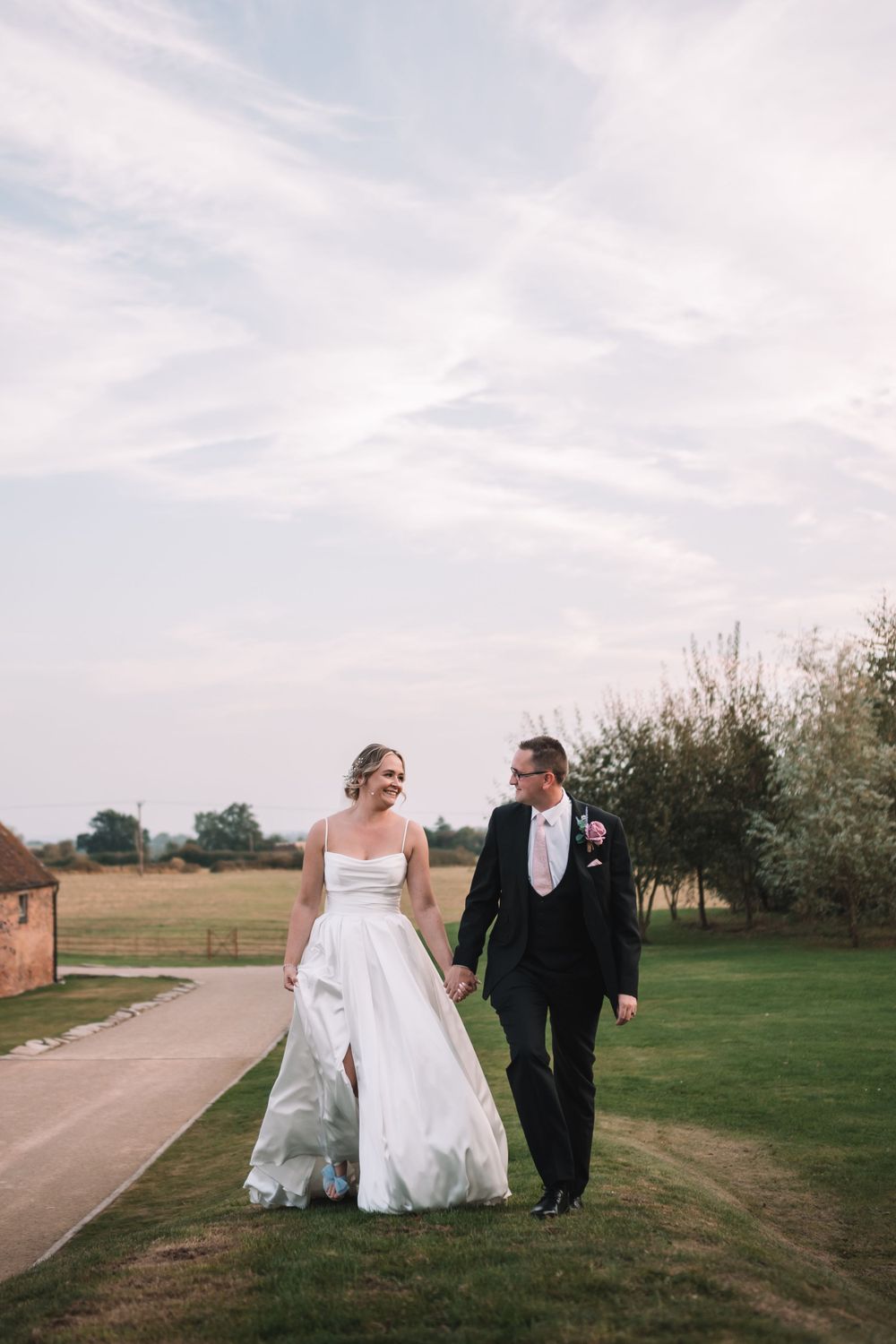 A couple walks hand in hand along a countryside path, the bride wearing a flowing white gown and groom in dark suit.