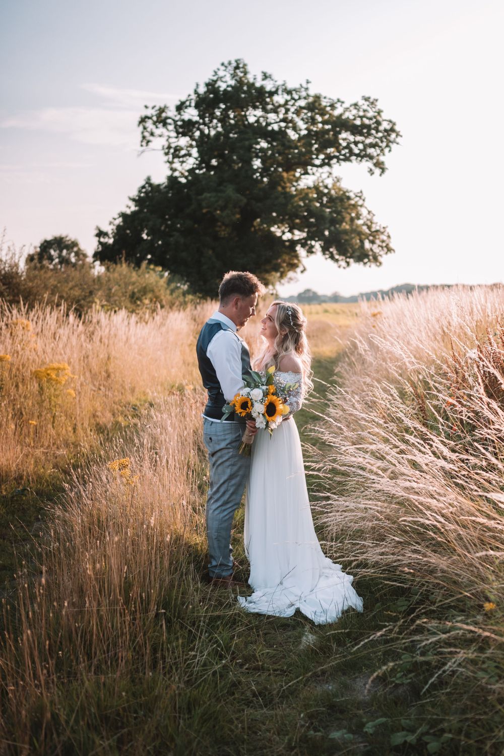 A couple embraces in a golden field of tall grass at sunset with a tree silhouetted in the background.