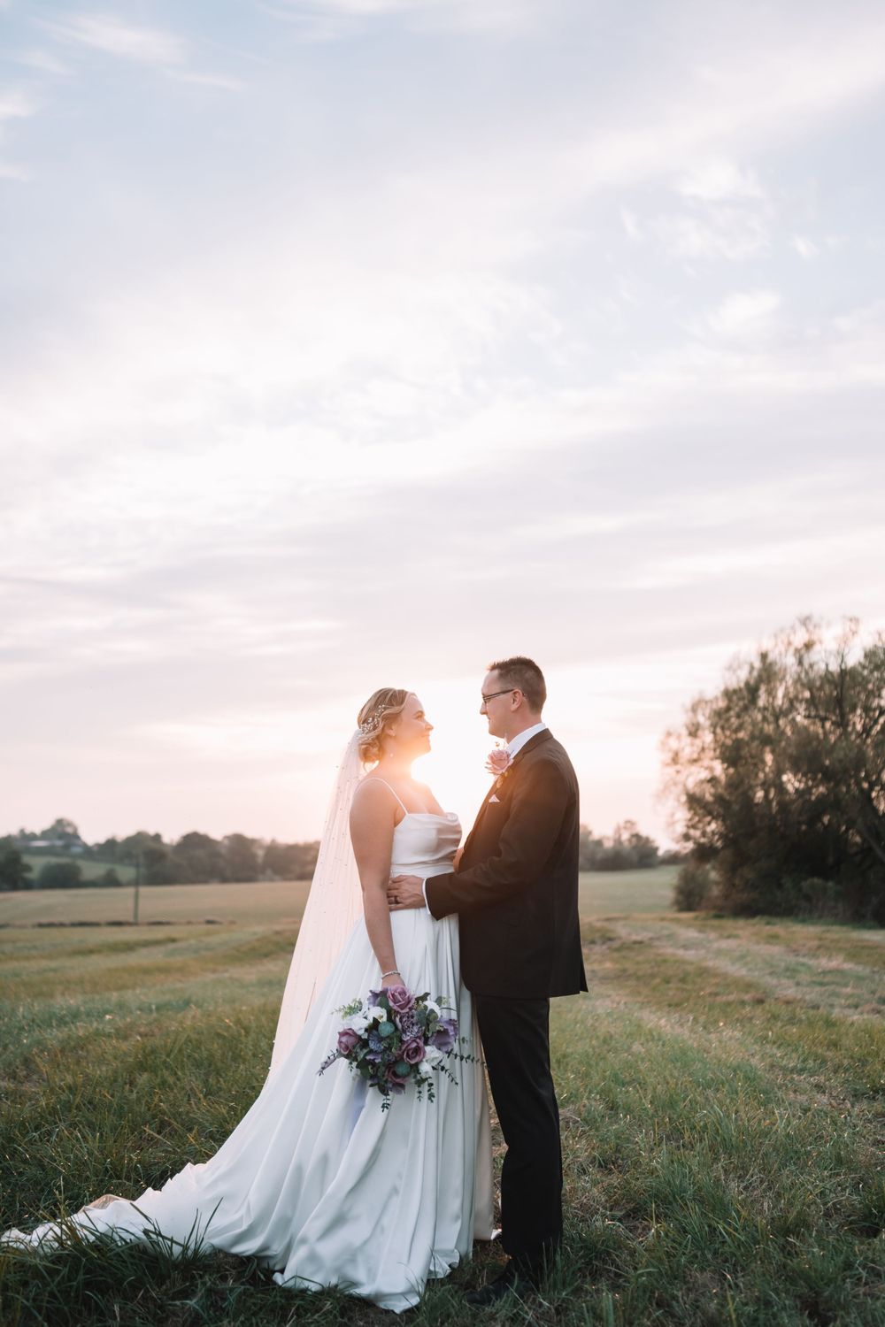 A bride and groom stand together in a grassy field during sunset, with a purple bouquet and flowing white dress.