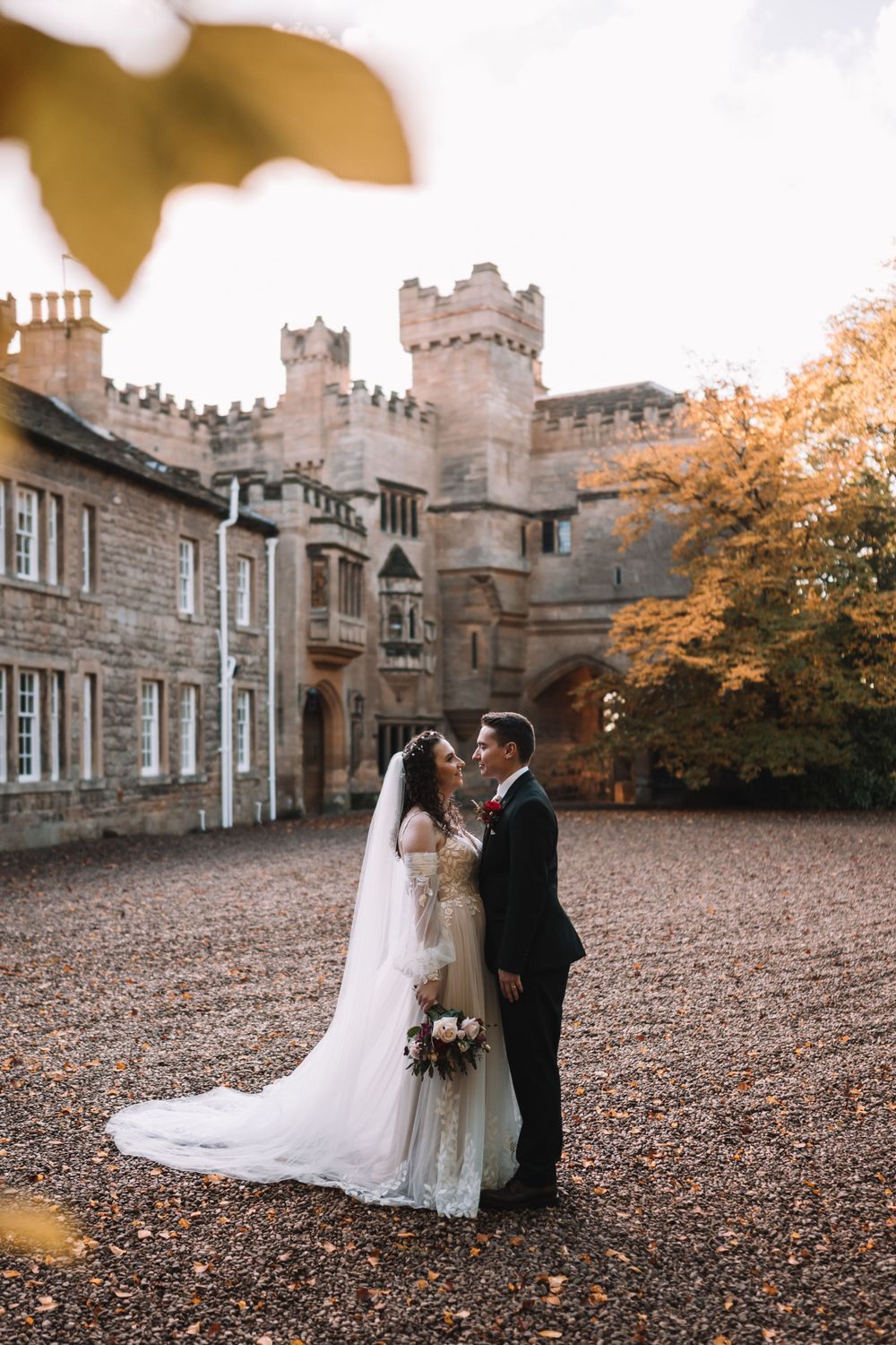 A wedding couple stands in front of a historic stone castle on an autumn day with fallen leaves covering the ground.
