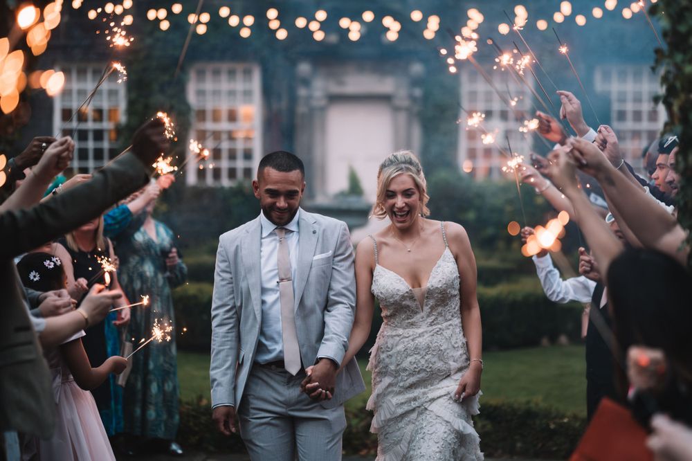 Newlyweds walk through guest sparkler exit at twilight outdoor wedding reception with string lights overhead.