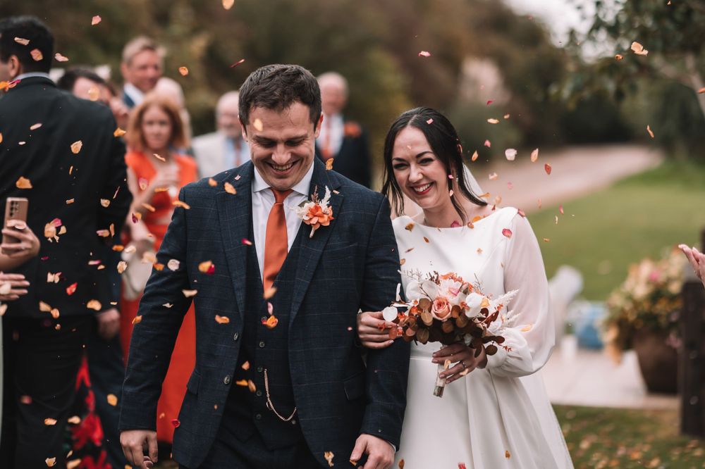 Couple walks down outdoor path amidst falling confetti after wedding ceremony as guests celebrate behind them.