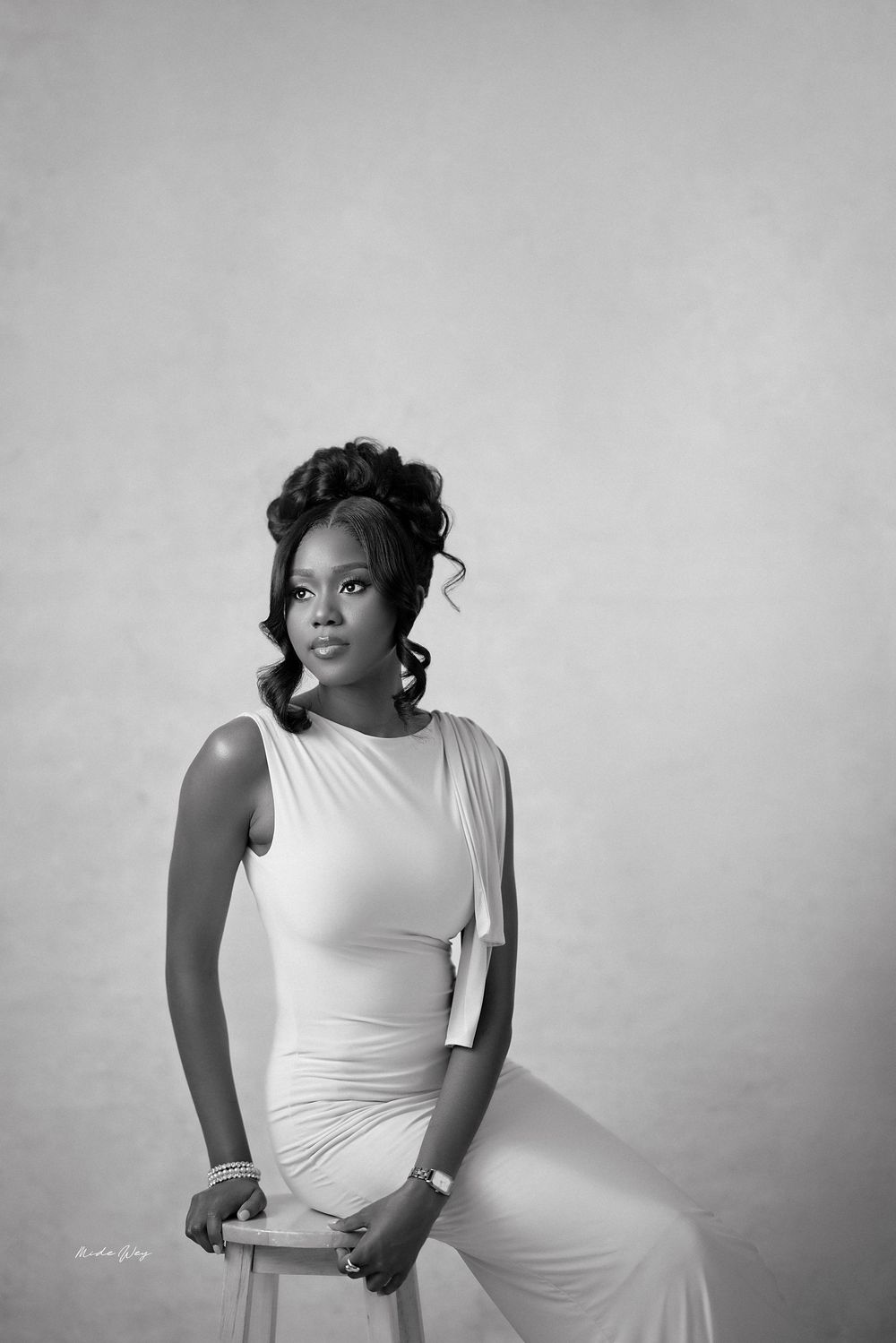 A black and white portrait of someone in an elegant white sleeveless dress seated gracefully against a plain background.