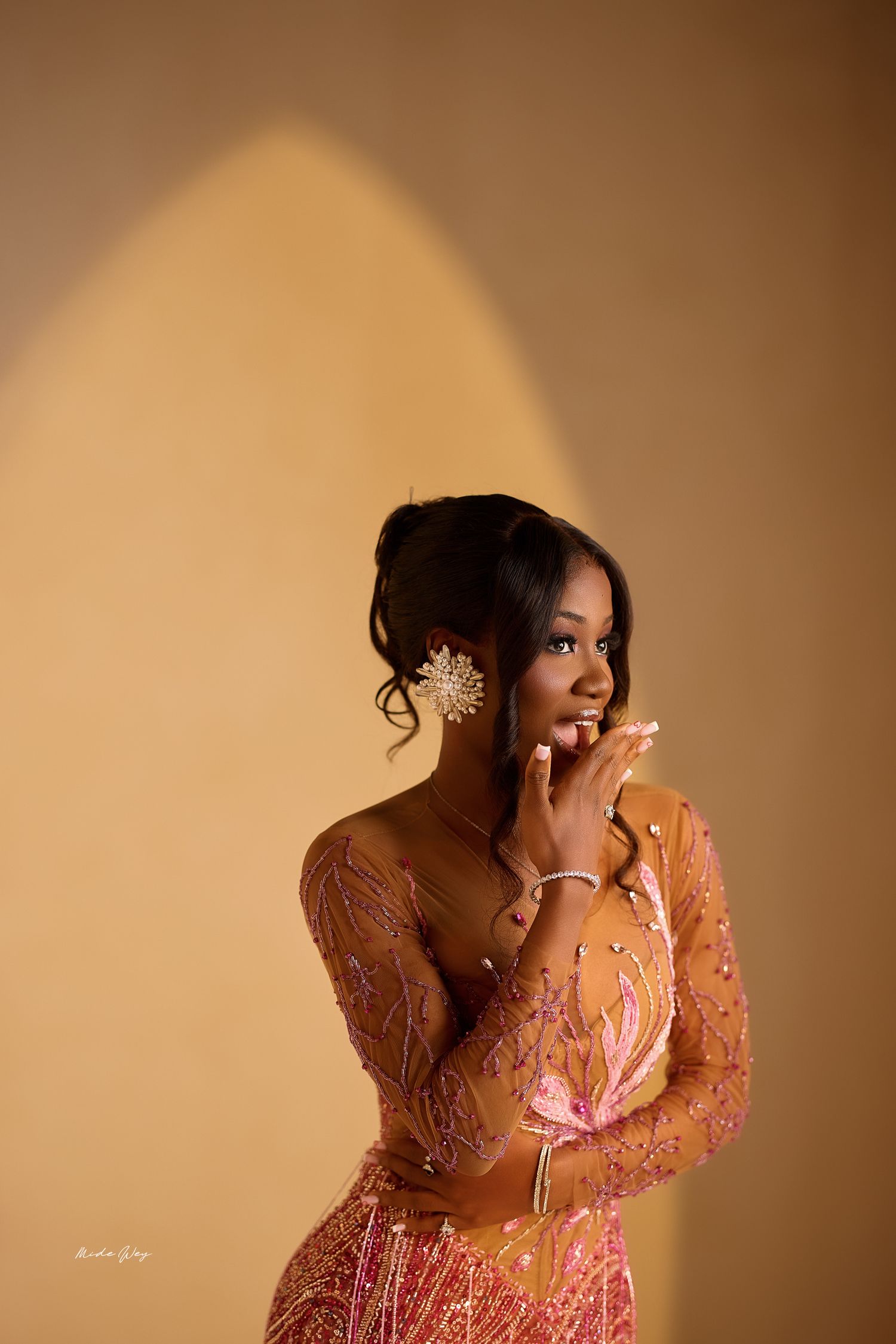 A woman in a pink embellished dress poses elegantly against a warm brown background in a glamour photoshoot.