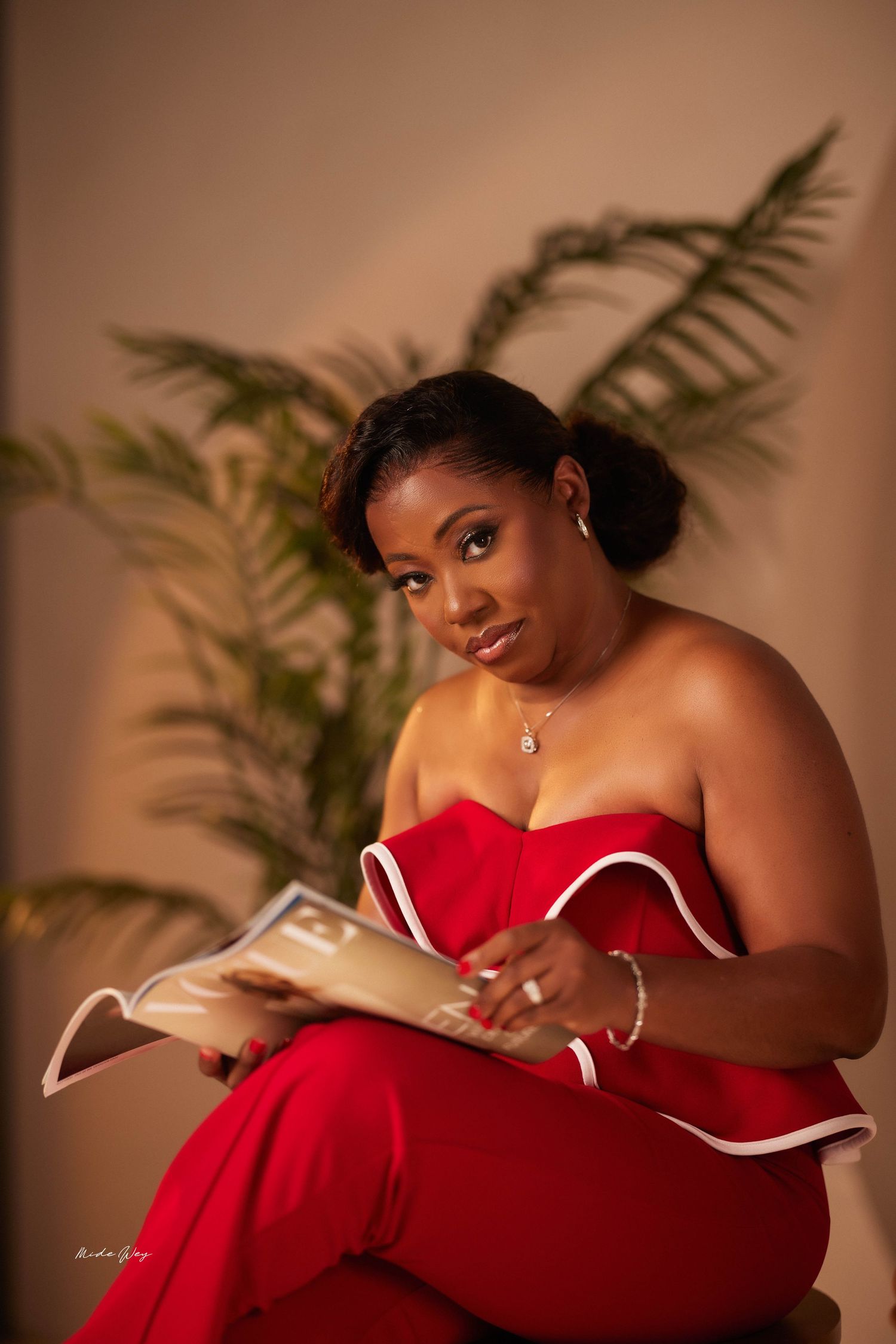 Woman in red dress poses gracefully while reading book in warm-toned studio setting with ferns.