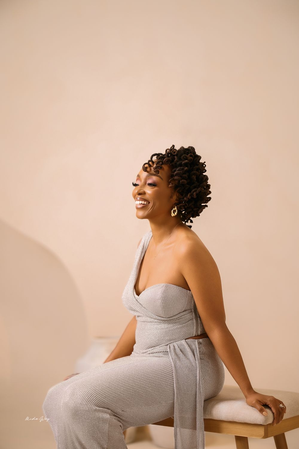 A pregnant woman in a grey dress laughs joyfully during a maternity photoshoot against a beige background.