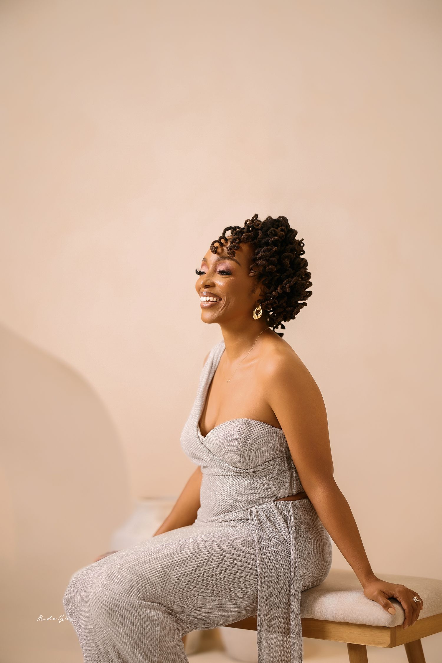 A pregnant woman in a grey dress laughs joyfully during a maternity photoshoot against a beige background.
