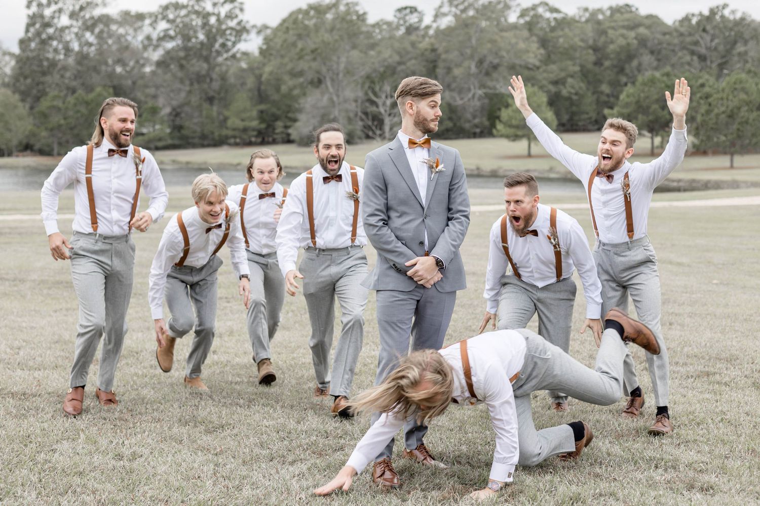Groomsmen in gray suspenders playfully pose while jumping in a field.