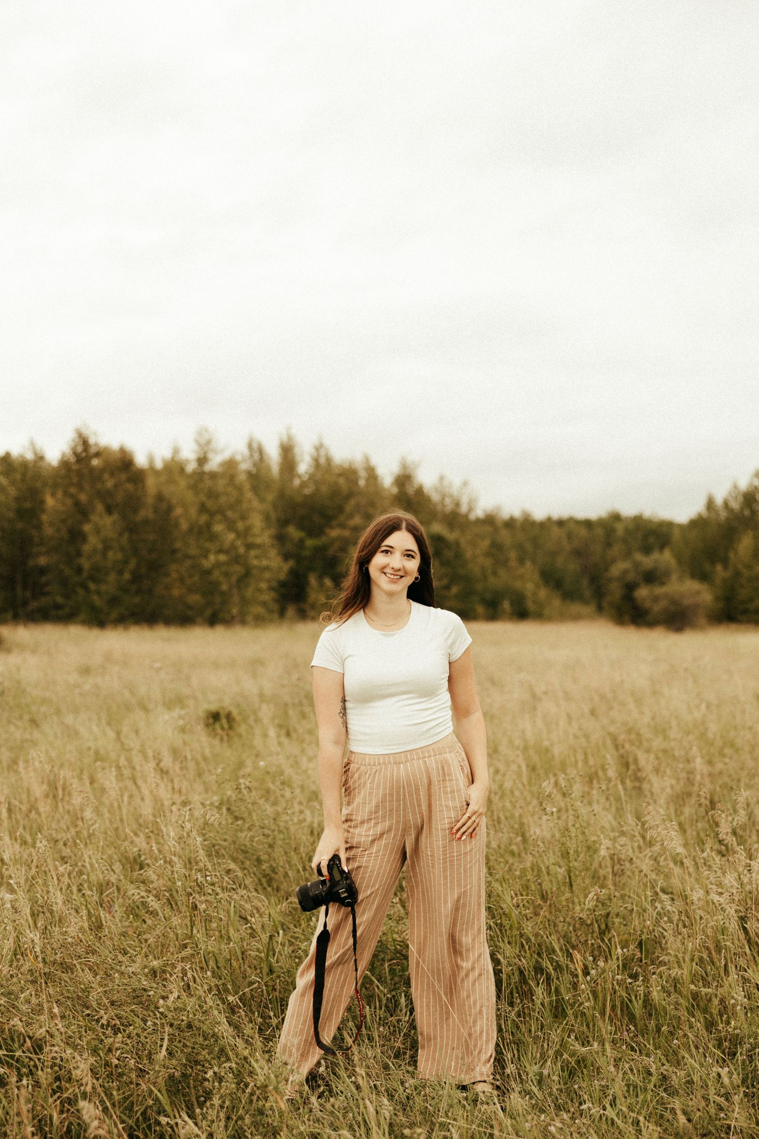 Brunette girl standing outside looking at the camera while holding her camera down by her side.