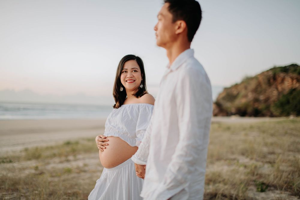 Pregnant woman in white off-shoulder dress smiles at man on beach during golden hour maternity photoshoot.