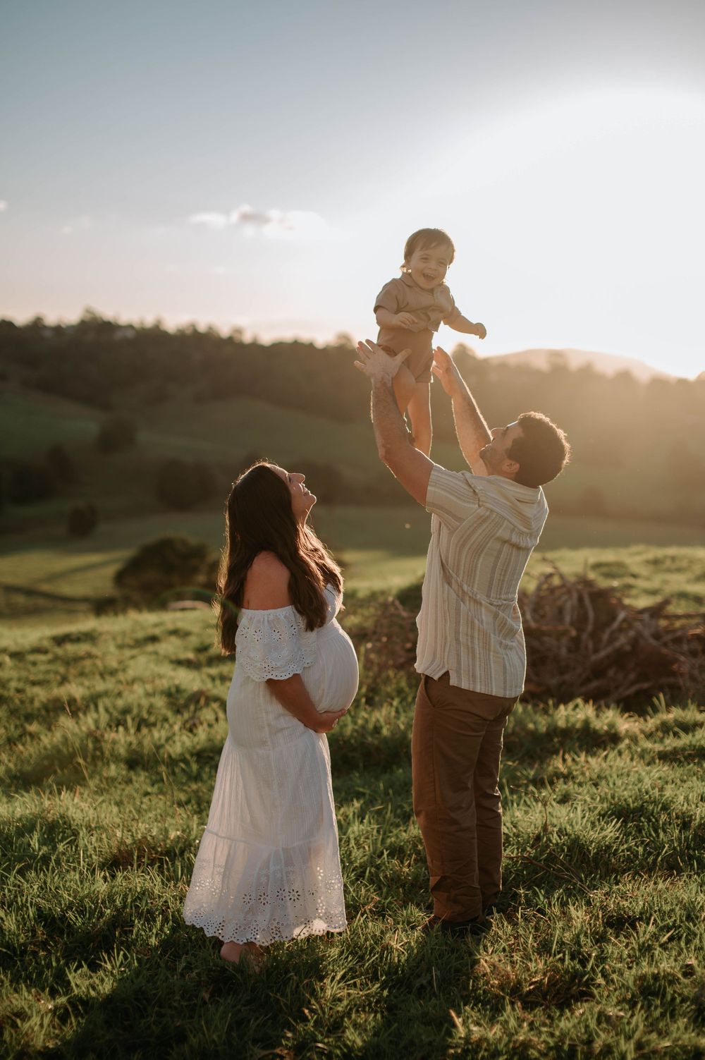 Pregnant woman in white dress and man lifting baby outdoors at golden hour on green hillside.
