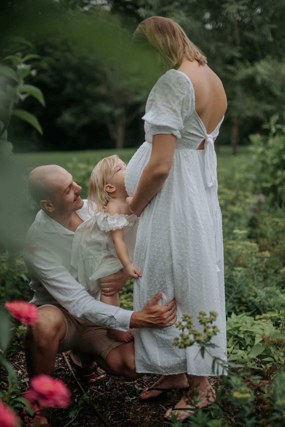 A bald man kneels in a garden beside a toddler and a pregnant woman in a white backless dress.
