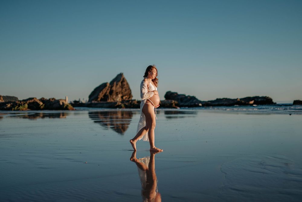 Pregnant woman in white shirt walks barefoot on reflective wet beach at golden hour with rocky coastline backdrop.