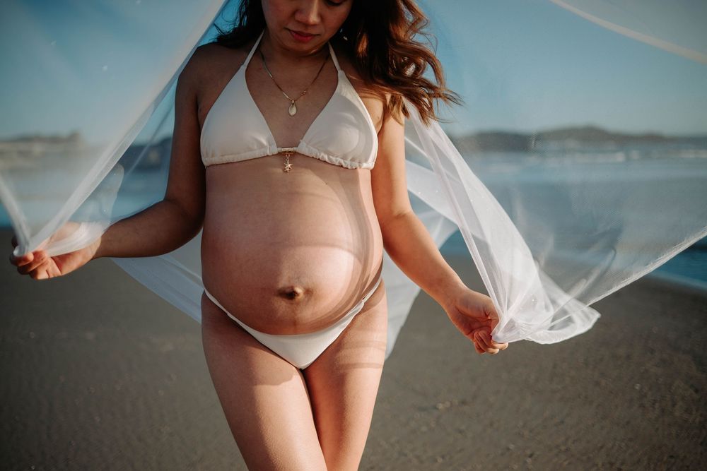 Pregnant woman in white bikini at beach holding flowing sheer fabric, showcasing baby bump during maternity photoshoot.