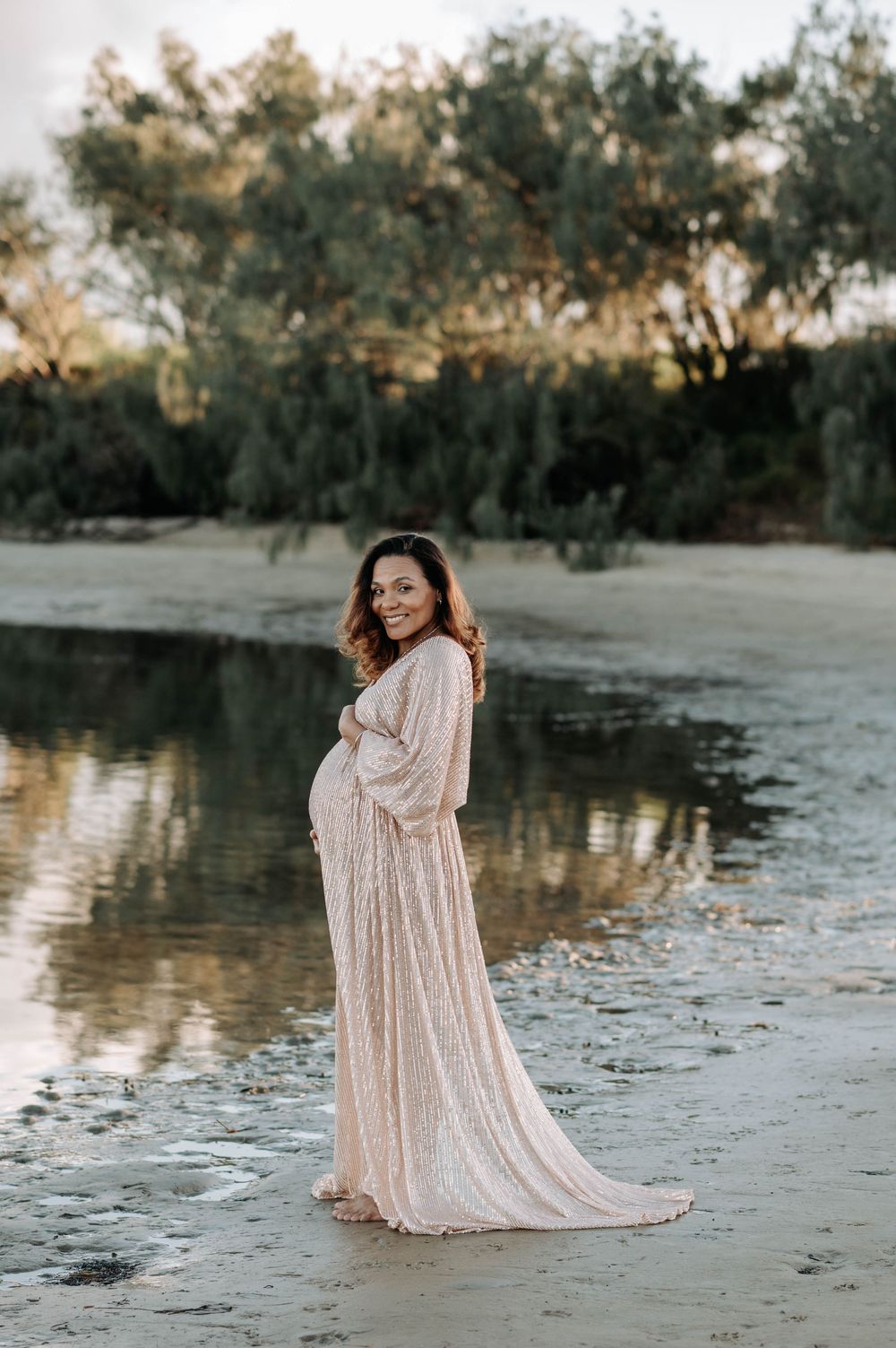 Pregnant woman in elegant sequined blush gown standing by a reflective lake shore with trees in background.