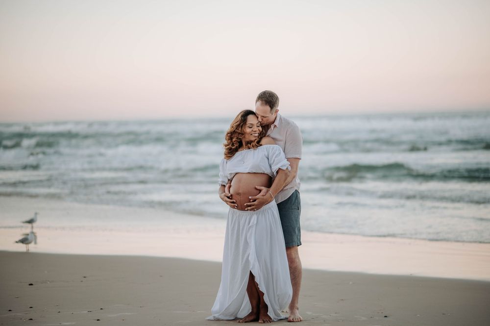 Couple sharing a tender maternity beach moment at sunset, woman in white flowing dress cradling baby bump.