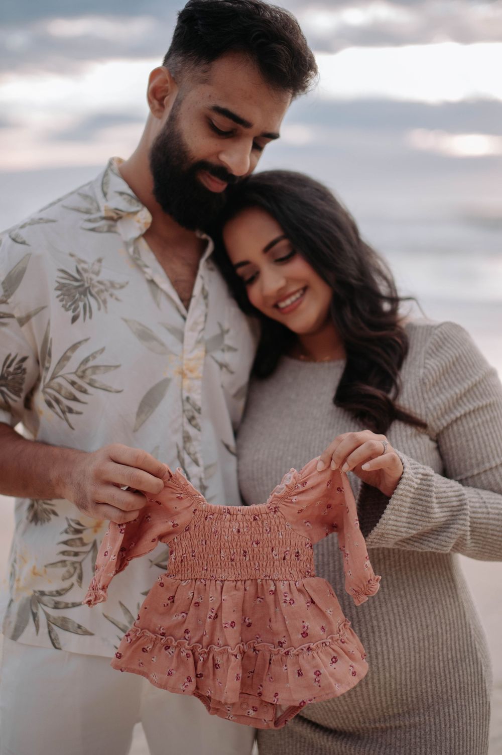 Couple holding a pink baby girl romper outdoors, announcing pregnancy in a tender maternity photo shoot.