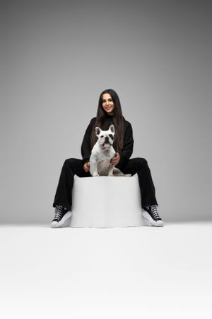 A French Bulldog sits with its owner on a white cylindrical platform against a gray background.
