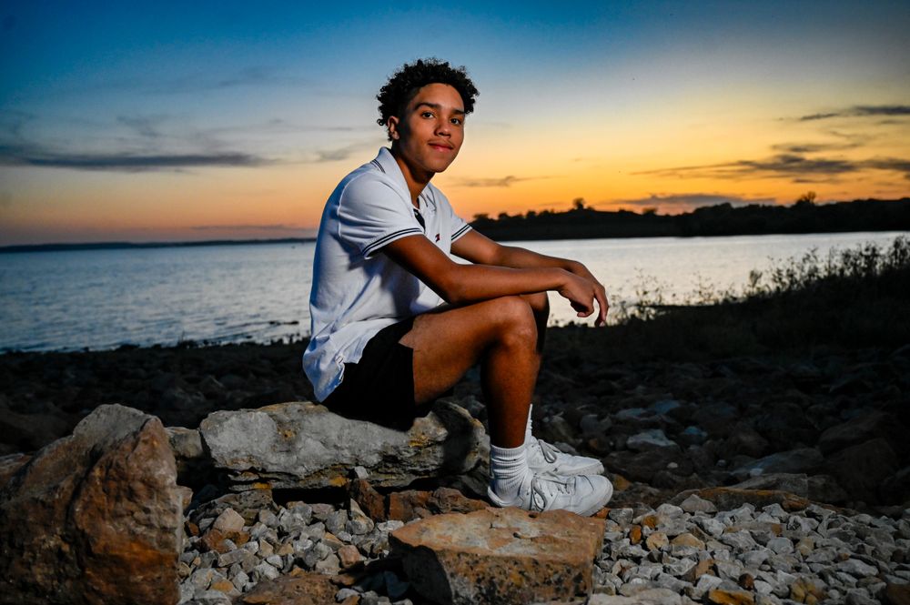 Person in casual attire sits on rocky shoreline during sunset.