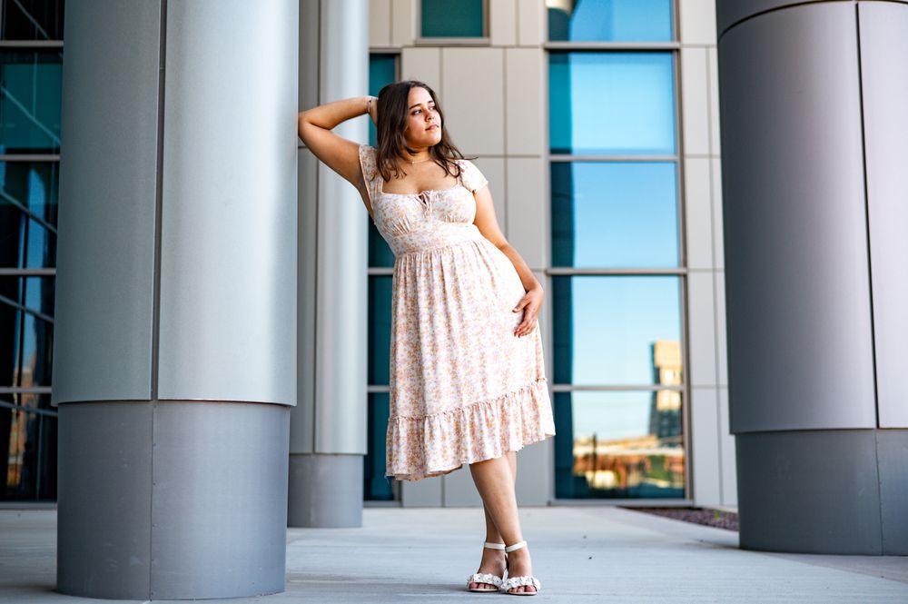 A woman in a light floral dress poses elegantly between modern architectural columns.