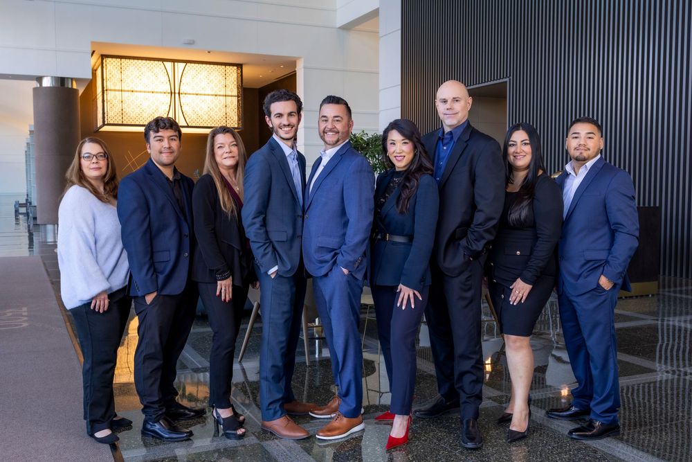 Group of business professionals in formal attire posing together in a modern office hallway.
