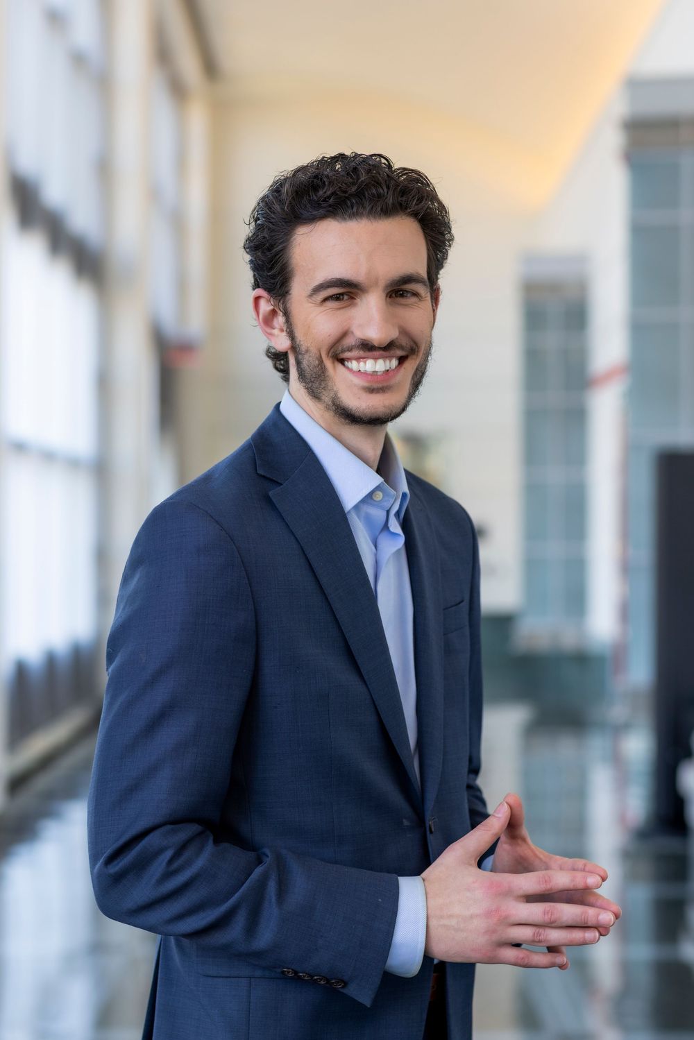 Professional in a navy suit and light blue shirt smiles warmly while gesturing in a modern office hallway.
