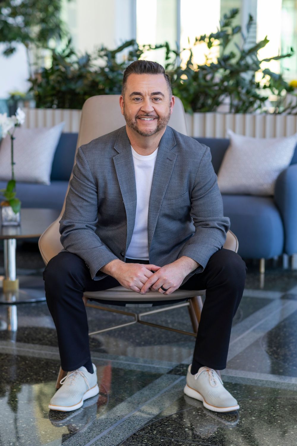 A person in a gray blazer and white t-shirt sits casually on outdoor steps while smiling at the camera.