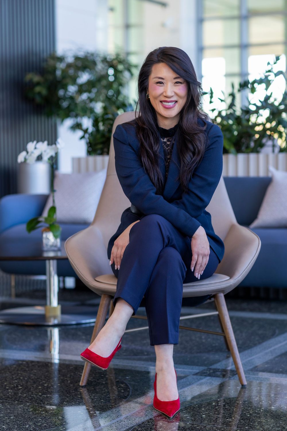 Professional woman in navy business suit sits elegantly in modern grey chair wearing striking red heels.