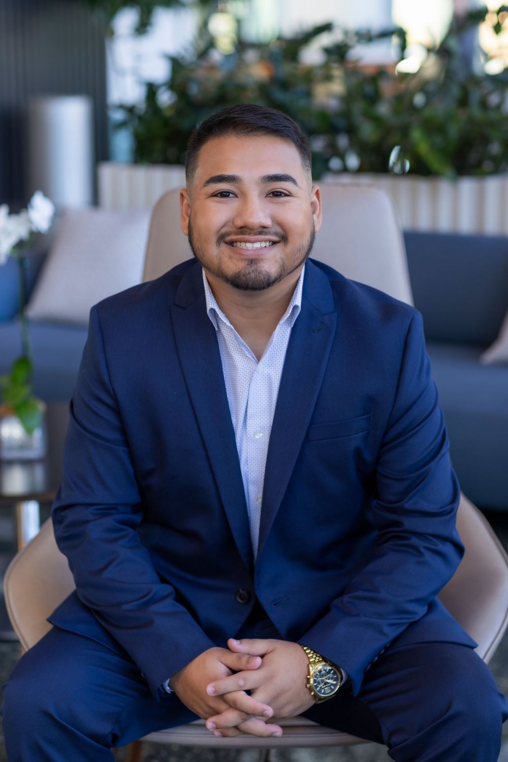 Professional wearing a navy suit and white dress shirt sits in a modern office setting with a warm smile.