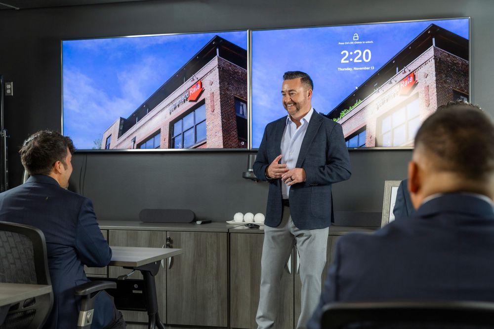 A presenter gives a business presentation in a modern conference room with multiple screens showing data.