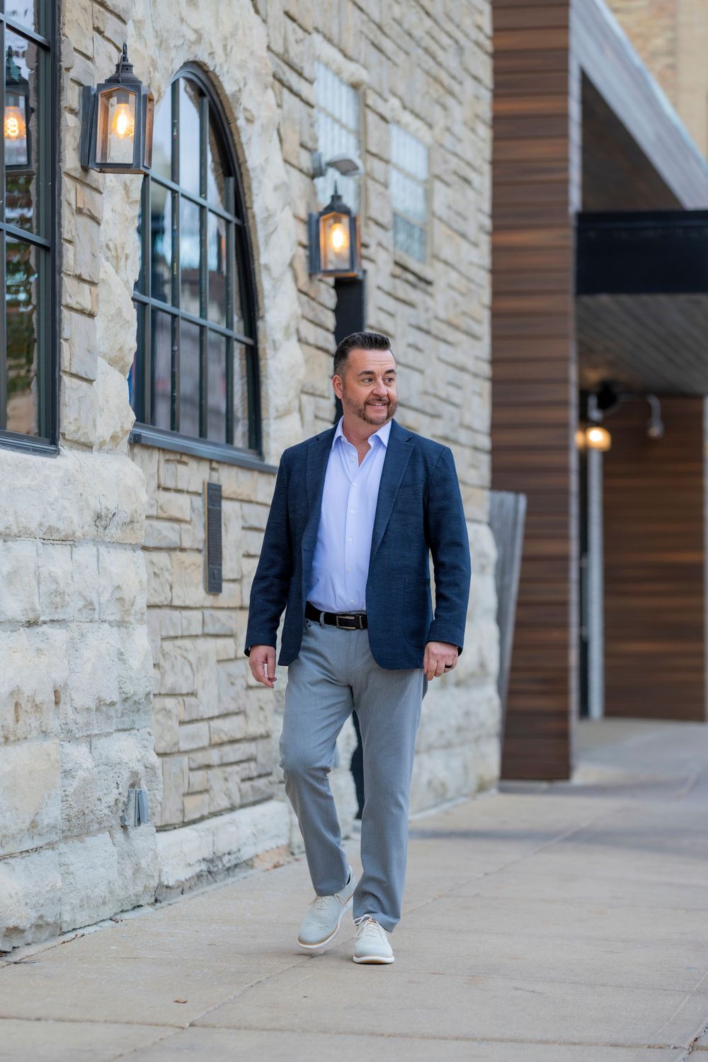 Business professional in navy blazer and grey pants walks confidently along stone building exterior.
