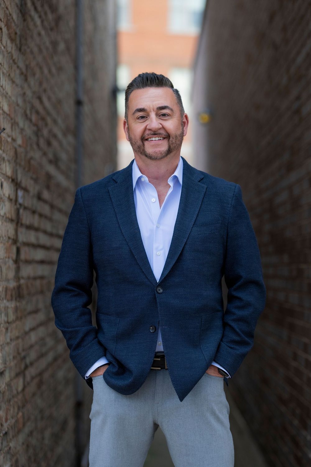 Professional headshot portraits of someone in a navy blazer and white shirt against a brick wall background.