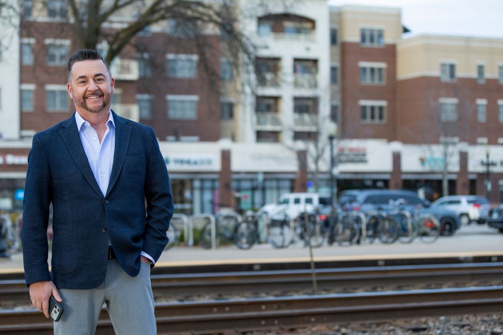 Professional in a navy blazer and dress shirt stands near train tracks with urban buildings in the background.