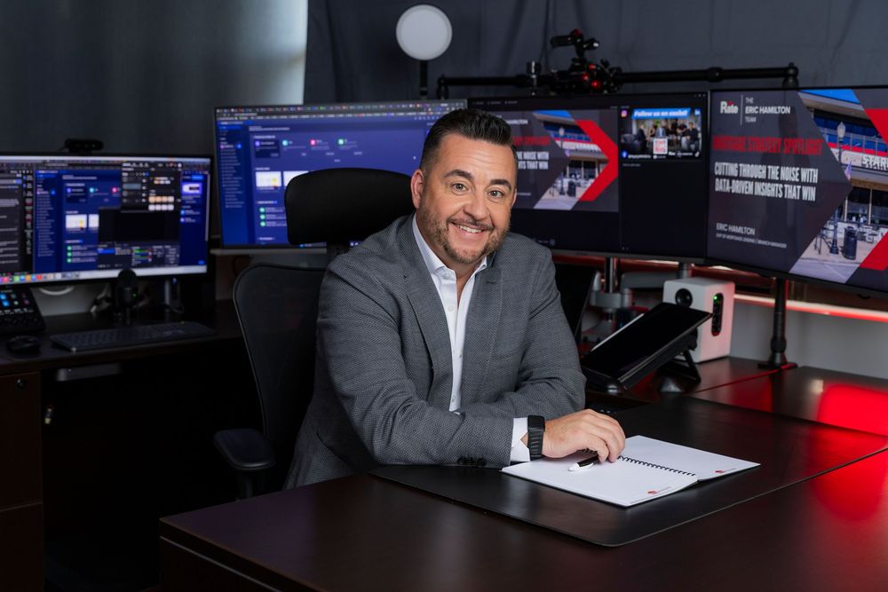 A person sits at a broadcast desk in a television news studio with multiple monitors and red lighting visible.