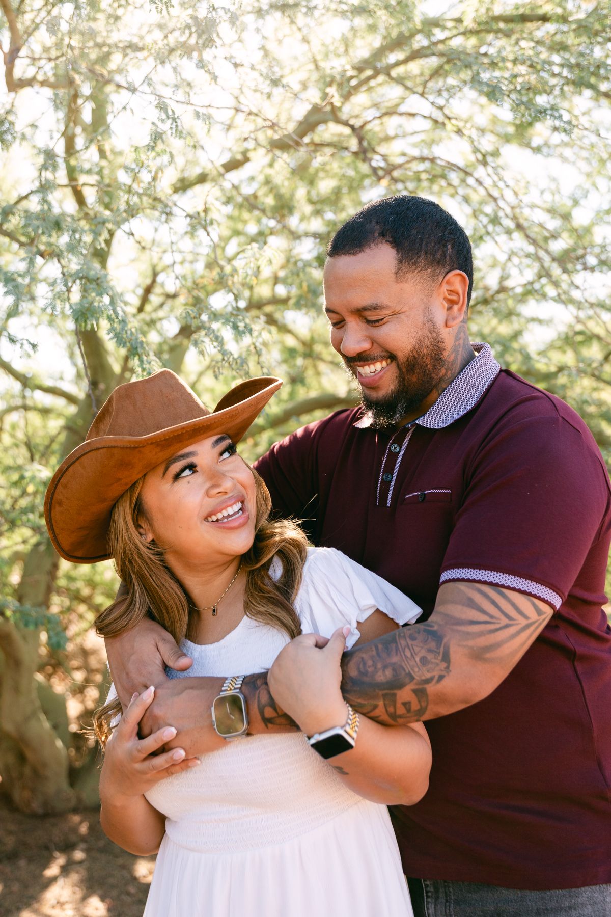 A man in a red shirt is hugging a woman in a white dress and a cowboy hat. They're smiling at each other with a green treen behind them