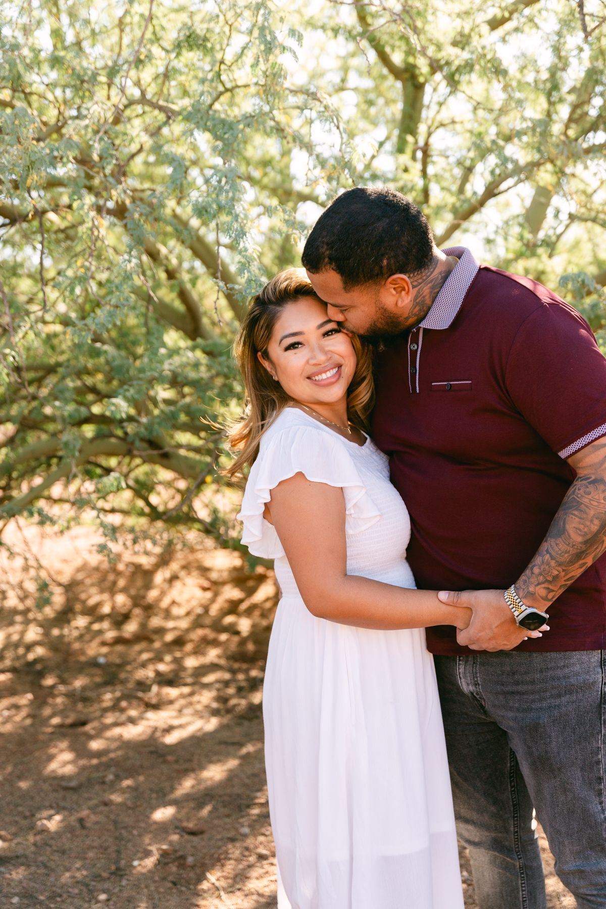 A man in a red shirt is kissing the forehead of a woman in a white dress. They're holding hands and are standing in front of a green tree.