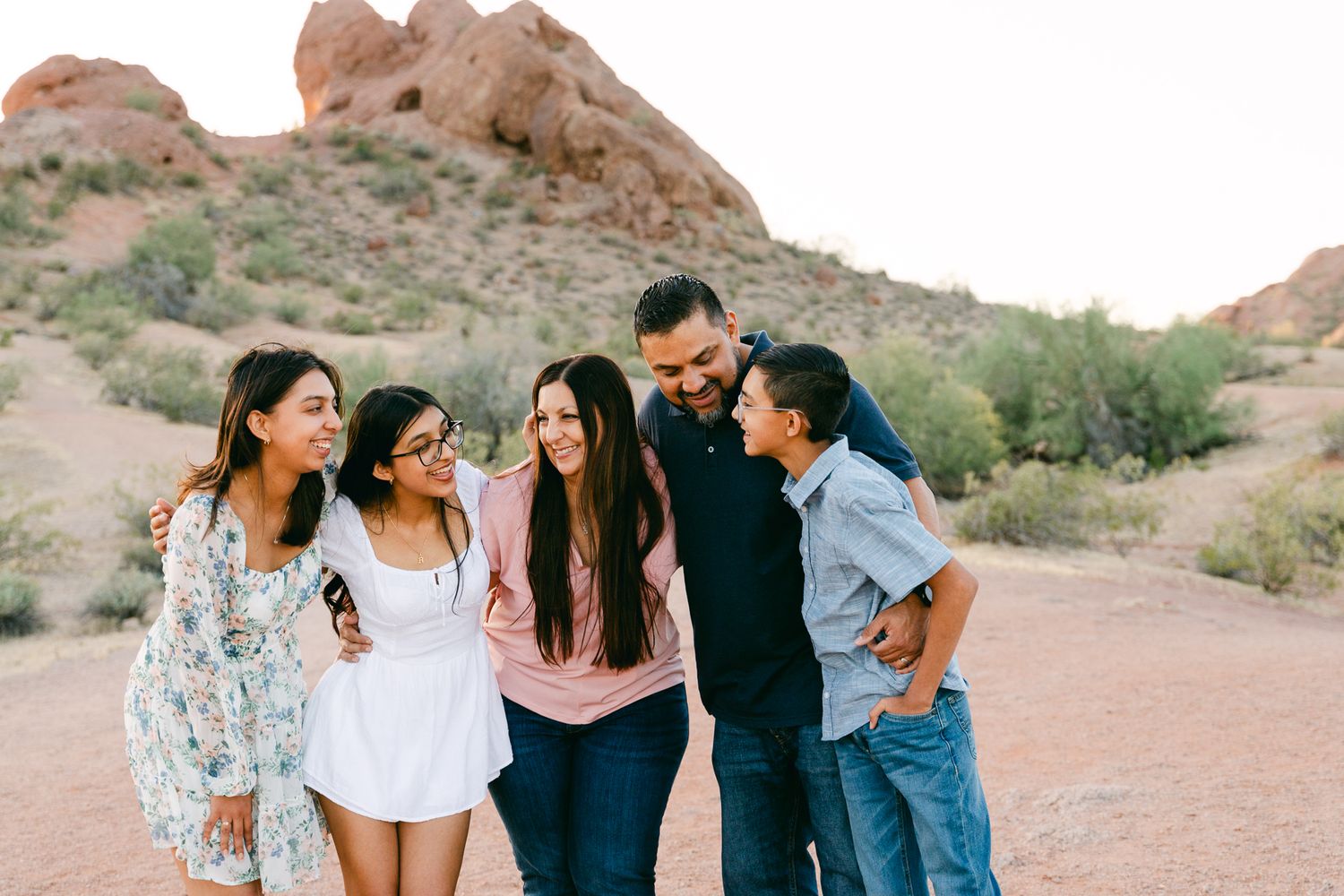 A family of five is laughing and looking around. There's two girls, a boy, a woman and man. There's greenery and a brown mountain behind