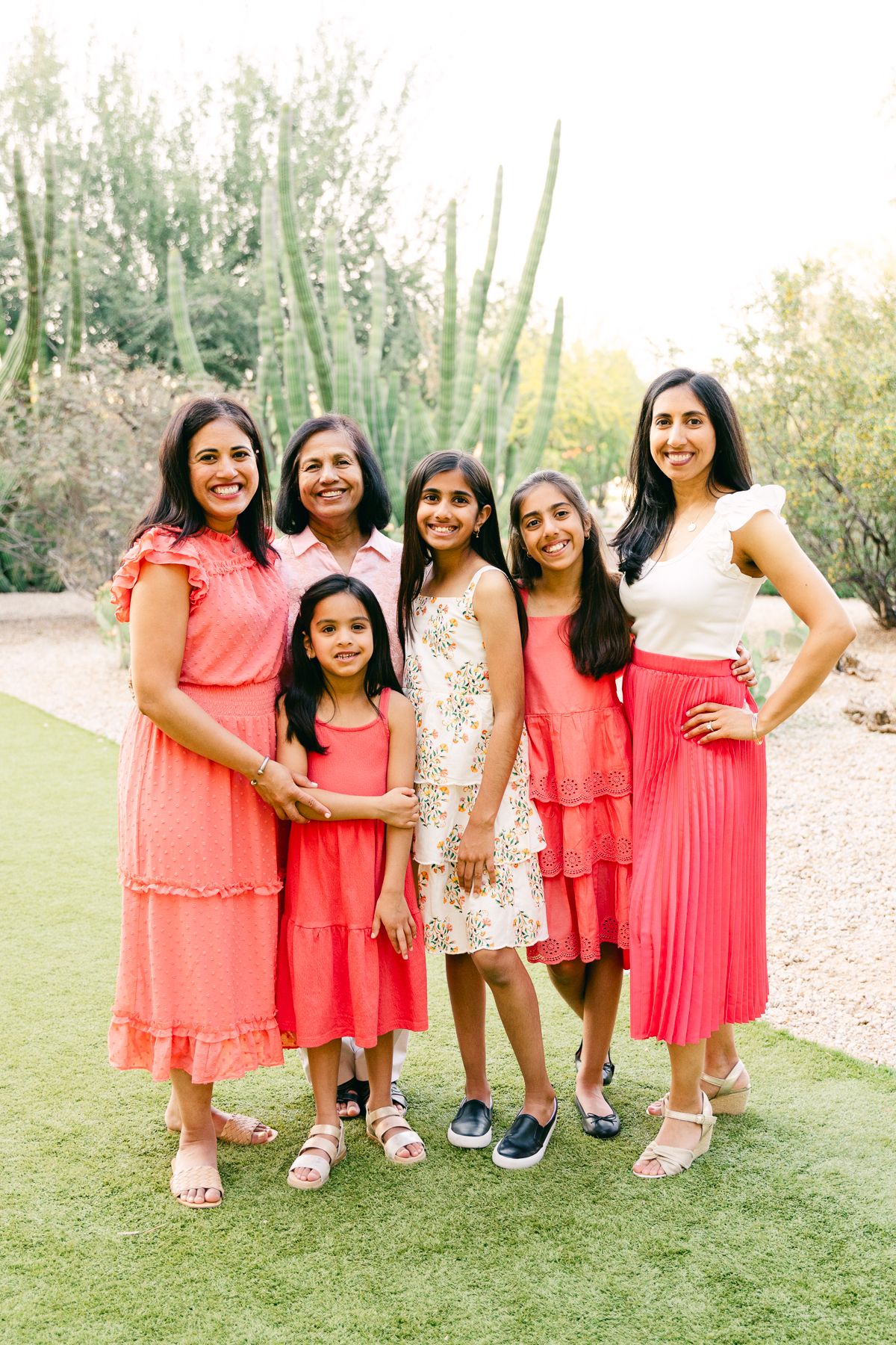 Three woman and 3 girls are standing toghether and smiling. They're in pink and white and there's cactus and desert plants behind them.