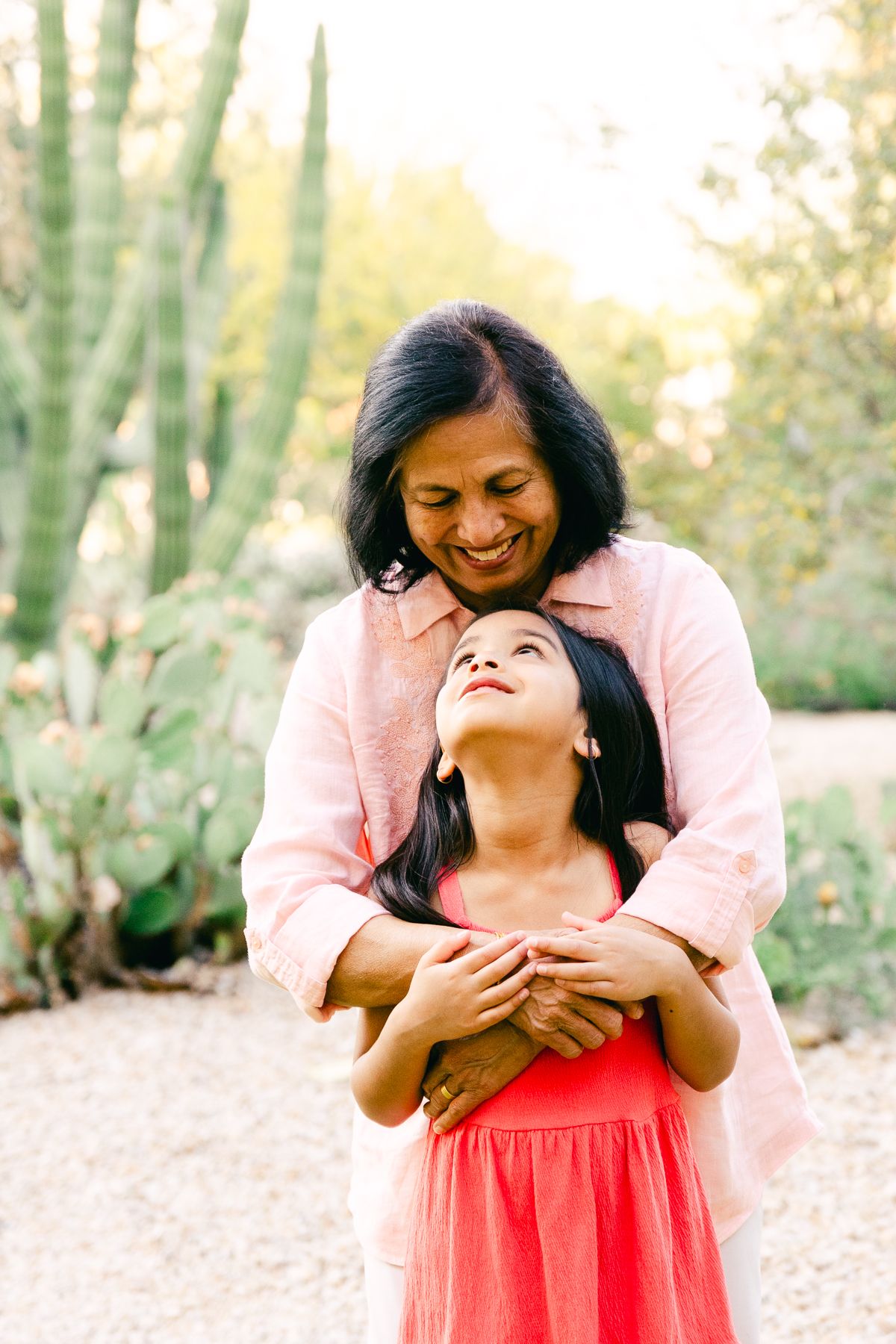 An older woman is hugging a younger girl and they're looking at one another. They're both wearing pink and there's dirt and cactus.