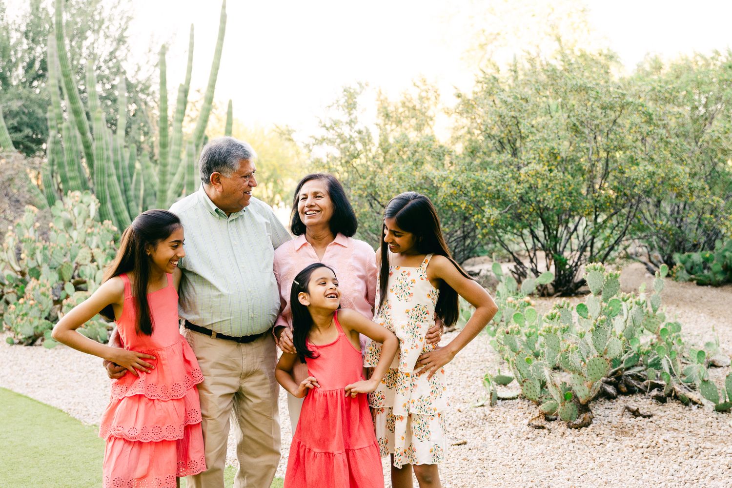 An older man, older woman and 3 girls are smiling and looking around. They're in green, pink and white and are standing in front of cactus.