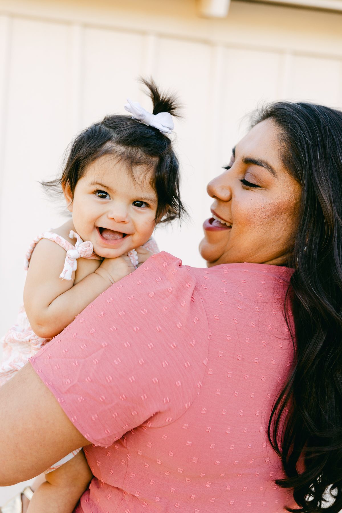 A woman in pink is holding a baby in pink. The baby has a white bow in her hair and smiling at the camera.
