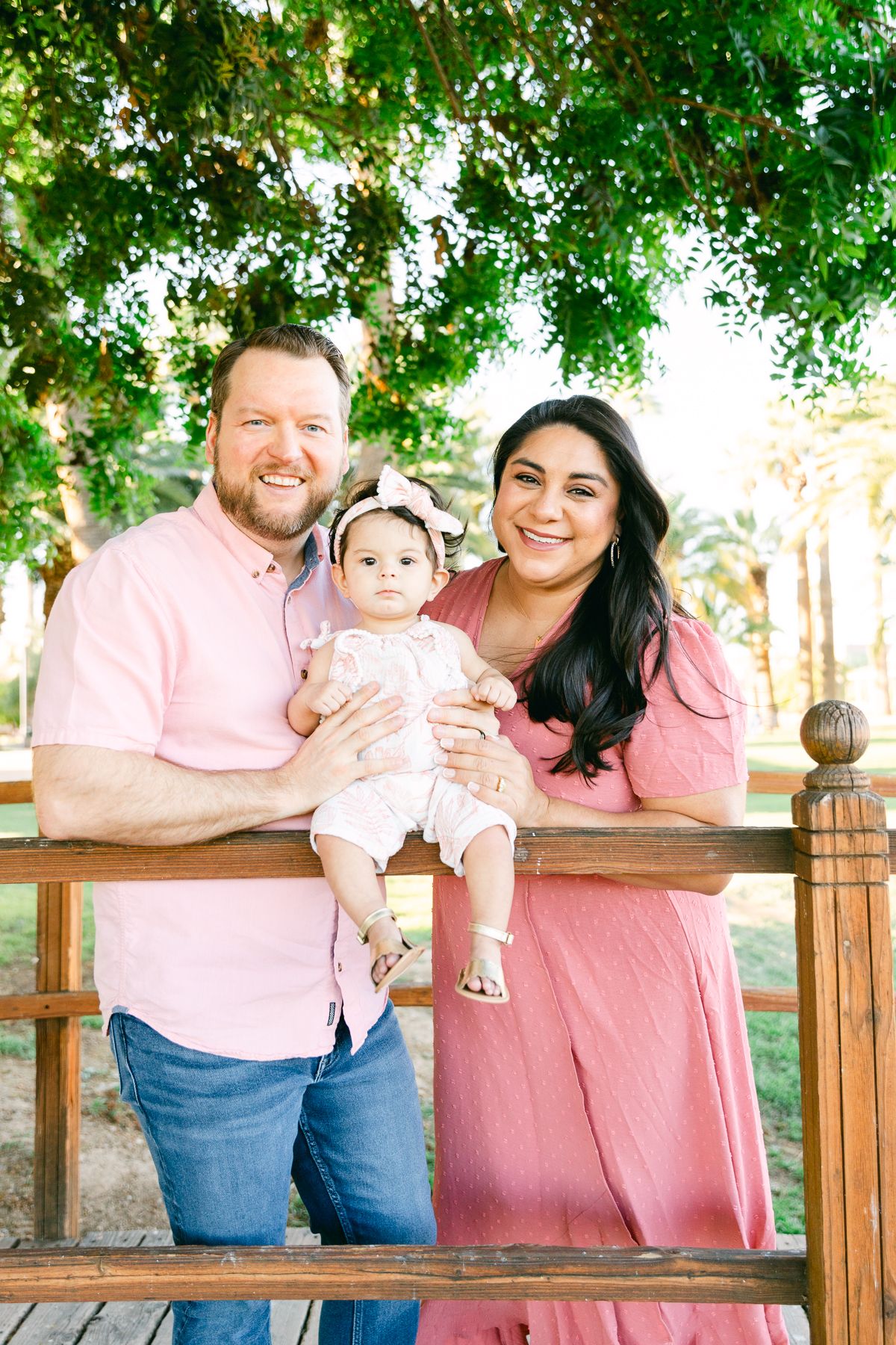 A man and woman are holding a baby. They're all in pink and are standing on a bridge with a tree overhead.